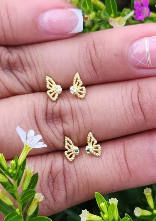 Primer plano de una mano con uñas cuidadas sosteniendo dos pares de aretes de mariposa de oro con piedras centrales verdes y transparentes, elaborados con Joyas en Oro Laminado de 18K, colocados en medio de follaje verde y flores blancas.