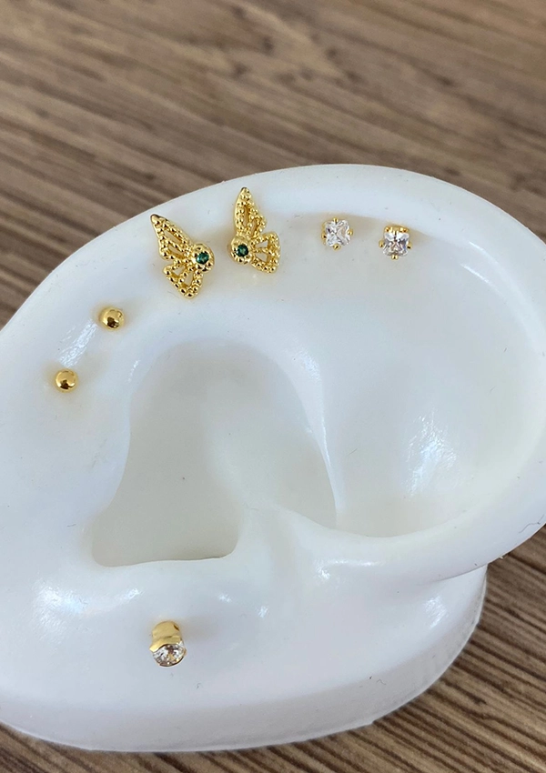 Un porta joyas blanco en forma de oreja muestra varios aretes, incluidos Joyas en Oro Laminado de 18K: aretes de mariposa de oro, pequeños aretes de botón de oro y un botón de oro con una sola piedra preciosa.