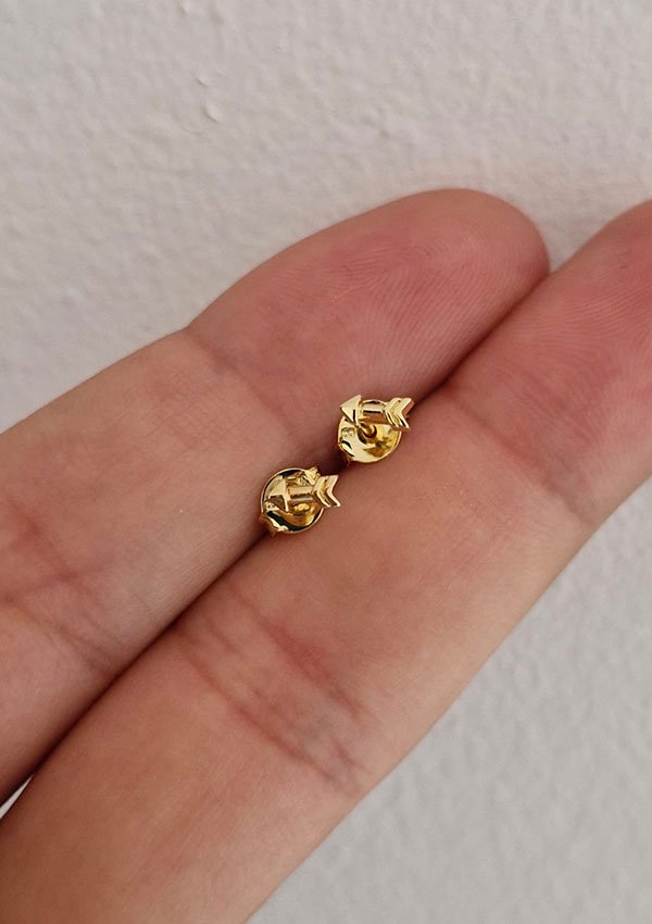 Una mano sostiene un par de pequeños aretes dorados en forma de gato elaborados con Joyas en Oro Laminado de 18K sobre un fondo neutro.