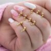 Una mano con uñas cuidadas, que combina esmalte blanco y nail art floral, sostiene varios pequeños aretes de estrellas de oro de la colección Joyas en Oro Laminado 18K.