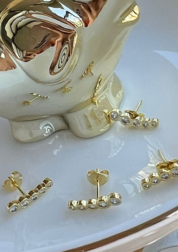 Un conjunto de aretes de joyas en oro laminado de 18k con cinco cuentas cada uno está dispuesto sobre una superficie blanca, con un objeto de cerámica reflectante en el fondo.