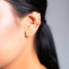 Primer plano de la oreja de una persona adornada con joyas de oro laminado de 18k, incluidos dos aros de oro en la hélice y cinco pequeños pendientes de oro en el lóbulo. El cabello largo y oscuro está atado hacia atrás, lo que resalta la elegante joyería de la oreja.