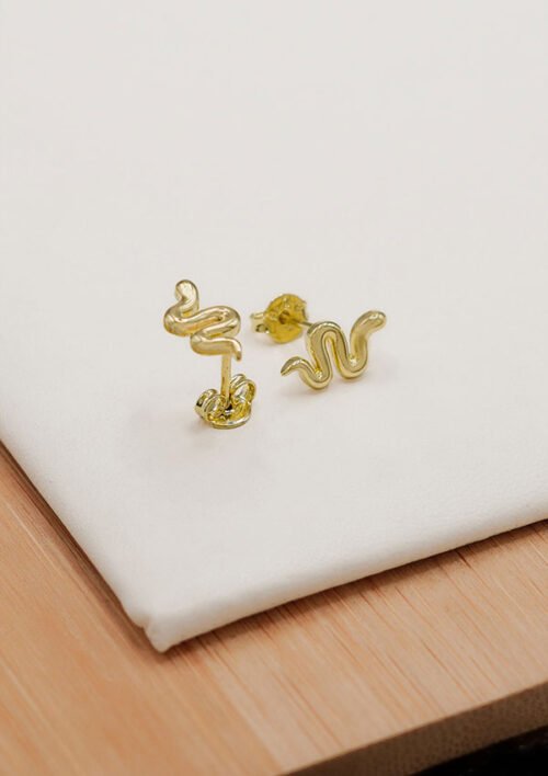 Presentamos los Aretes Snake, aretes de oro con forma de serpiente elaborados con oro laminado de 18 quilates, exhibidos sobre una tela blanca sobre una superficie de madera. Perfecto para añadir un toque elegante a tu colección de joyas.