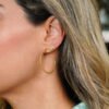 Primer plano de la oreja de una persona adornada con joyas, incluido un arete de aro Aretes Snake en oro laminado de 18 quilates y un pequeño ear cuff de oro en forma de ola. La persona tiene cabello rubio y lleva una blusa verde.