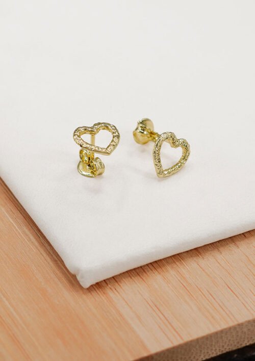 Aretes Little Heart, pendientes en forma de corazón con diseño texturizado, elaborados en oro laminado de 18K, colocados sobre una tela blanca sobre una superficie de madera.