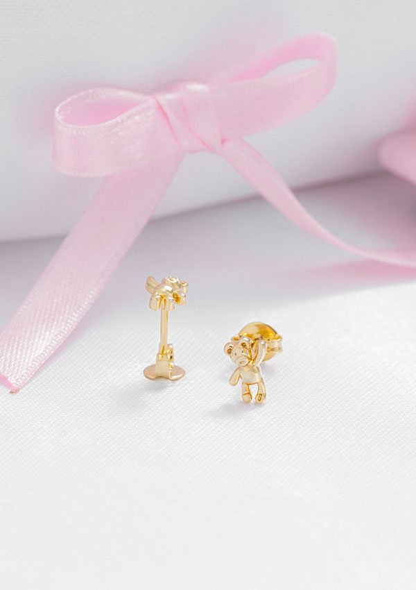 Los Aretes Osito, unos pendientes dorados con forma de osito fabricados en Oro laminado 18K, se colocan sobre una superficie blanca con un lazo rosa de fondo, luciendo joyas elegantes para cualquier ocasión.