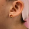 Primer plano de la oreja de una persona que lleva un Aretes Rainbow, un pequeño arete circular con cuentas multicolores elaborado con joyas y Oro laminado de 18 quilates, con una cabeza de tela rosa que cubre ligeramente visible.