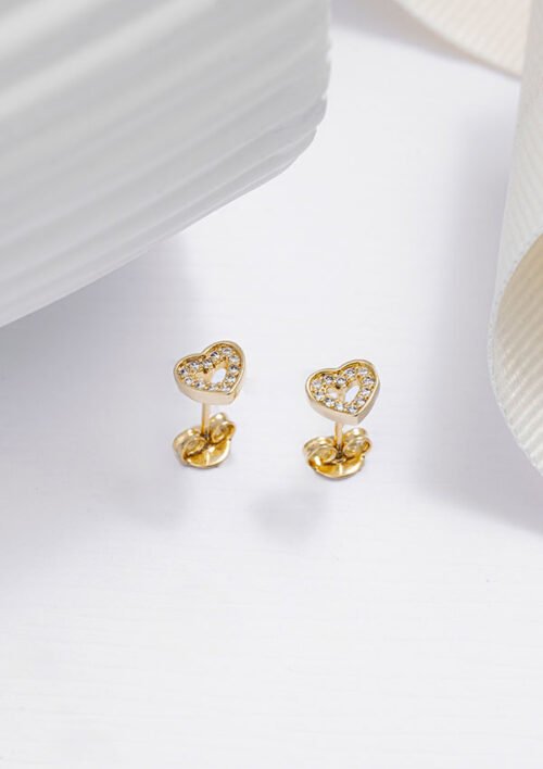 Primer plano de un par de Aretes Baby Love, con aretes de oro en forma de corazón adornados con pequeños cristales, elaborados en Oro laminado de 18K, elegantemente colocados sobre una superficie blanca con objetos blancos texturizados en el fondo.