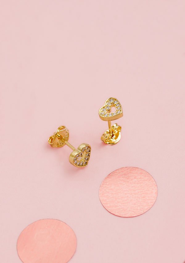 Aretes Baby Love, pendientes de oro en forma de corazón adornados con pequeños cristales incrustados, expuestos sobre un fondo rosa con adornos circulares de color rosa. Estas exquisitas joyas hechas de Oro laminado 18K añaden un toque de elegancia y sofisticación a cualquier atuendo.