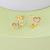 Los Aretes Double Free Love son unos aretes en forma de corazón de oro laminado de 18 quilates adornados con piedras preciosas multicolores en un diseño de corazón abierto, bellamente presentados sobre una superficie de color verde pastel y violeta.