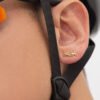 Primer plano de la oreja de una persona adornada con un pendiente Aretes Bicycle elaborado en Oro laminado 18K, con una correa de casco negra que cruza delicadamente sobre la oreja.