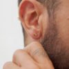 Primer plano de la oreja de una persona adornada con pequeños pendientes Aretes Bicycle oro laminado de 18K. La persona tiene el pelo corto, barba recortada y un dedo tatuado visible cerca de la oreja.