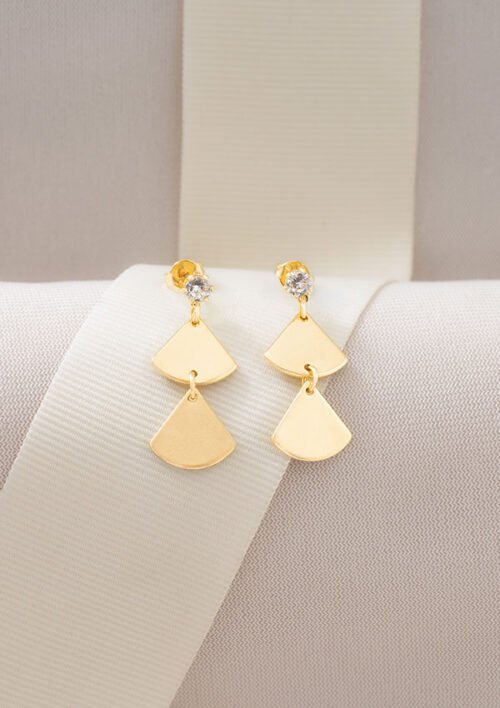 Aretes egipcios formados por unos pendientes geométricos de oro con dos formas triangulares y un pequeño diamante, elegantemente expuestos sobre una tela de color claro y con una cinta de fondo. Estas exquisitas piezas forman parte de nuestra colección de joyas de oro laminado de 18K.