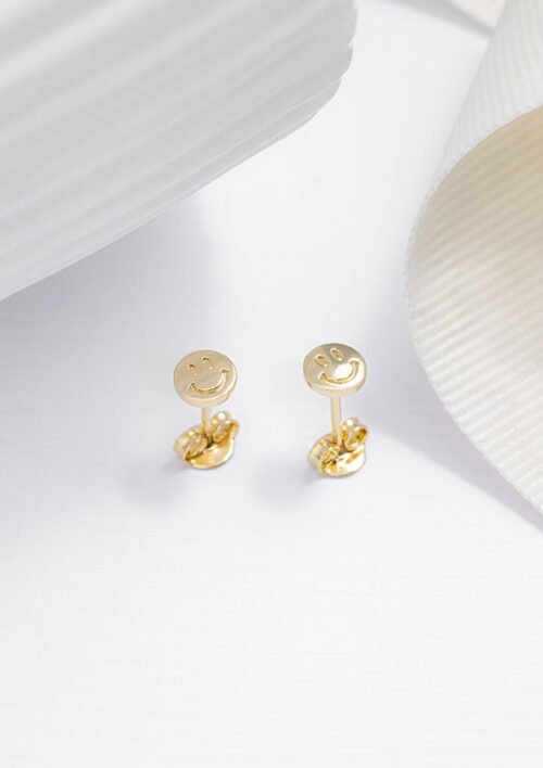 Aretes Smile, aretes de oro con diseño de carita sonriente, elaborados en oro laminado de 18K, elegantemente colocados sobre una superficie blanca con detalles texturizados en blanco y crema en el fondo. Estas exquisitas joyas añaden un toque de fantasía a cualquier conjunto.