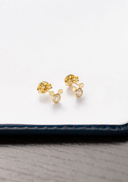 Un par de Aretes Mic Circonia con pequeños diamantes redondos y un diseño inspirado en Mickey Mouse, exhibidos sobre una superficie blanca y negra. Perfectas para añadir un toque de magia a cualquier conjunto, estas joyas son encantadoras y elegantes.