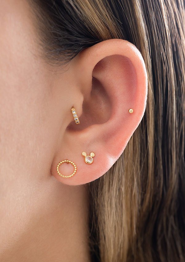 Primer plano de una oreja adornada con múltiples Aretes Mic Circonia: un pequeño pendiente de oro en el cartílago superior, un aro en el cartílago medio y tres pendientes de diferentes diseños en el lóbulo inferior, todos elaborados con el exquisito Oro laminado de 18K.