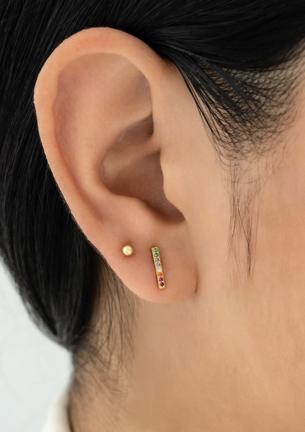 Primer plano de la oreja de una persona adornada con dos exquisitas piezas de la Línea Arco Iris de Aretes: un pequeño perno bañado en oro de 18 quilates en el lóbulo superior y un arete de barra vertical con piedras multicolores en el lóbulo inferior, ambos elaborados con un impresionante oro laminado de 18 quilates.