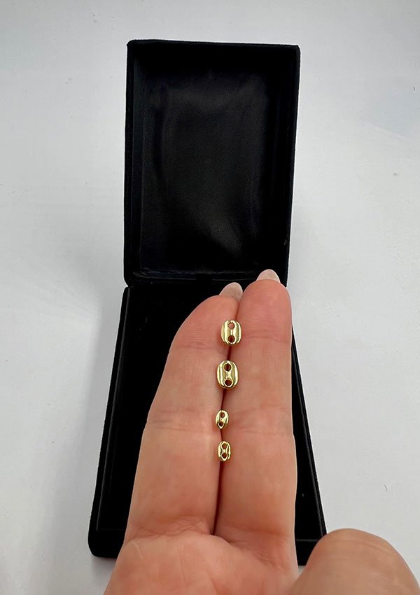 Una mano sostiene cuatro pequeños Aretes Gucci Finito, ovalados y de color dorado, frente a una caja negra abierta sobre una superficie blanca.