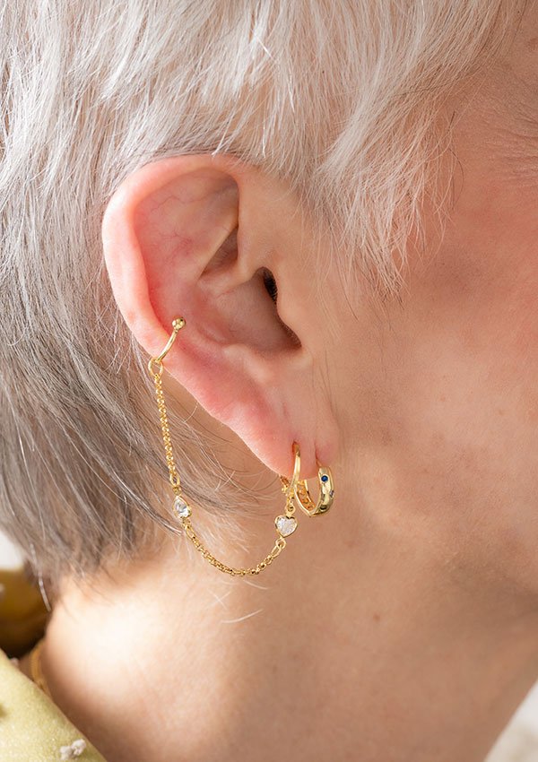 Primer plano de la oreja de un hombre adornada con las Candongas Sunset, un pendiente de cadena de oro laminado de 18K unido a una tachuela y un pequeño aro. La persona tiene el pelo corto y blanco, resaltando la elegancia de estas exquisitas joyas.