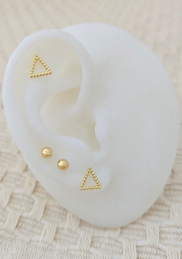 Una muestra de Aretes Triángulo París minimalista, con diseños triangulares y circulares, exhibidos sobre un modelo blanco con forma de oreja, colocado sobre una superficie texturizada de color crema. Estas joyas están elaboradas con Oro laminado de 18 quilates para brindar una elegancia duradera.