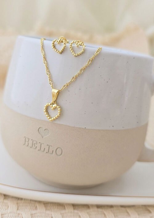 Un par de Aretes Corazón París y un collar con colgante de corazón de oro a juego, ambos elaborados en oro laminado de 18 quilates, exhibidos en una taza beige y blanca con "HOLA" grabado. Estas exquisitas joyas añaden un toque de elegancia a cualquier entorno.