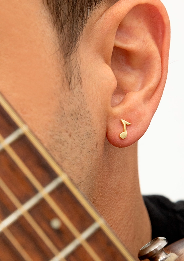 Primer plano de la oreja de una persona que lleva un pendiente de nota musical de oro, elaborado con Joyas en Oro Laminado de 18K, con el borde del mástil de una guitarra visible en primer plano.