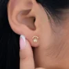 Primer plano de la oreja de una persona con un pequeño pendiente de oro en forma de corona. Los dedos de la persona, con esmalte de uñas rosa y blanco, tocan ligeramente la oreja. Esta elegante pieza forma parte de la colección Joyas en Oro Laminado 18K, que añade un toque de lujo al estilo cotidiano.