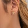 Vista de cerca de la oreja de una persona que lleva los "Aretes Sol redondo 8mm", que son pequeños aretes redondos de oro con diseño de rayos de sol, elaborados con joyas de oro laminado de 18K.