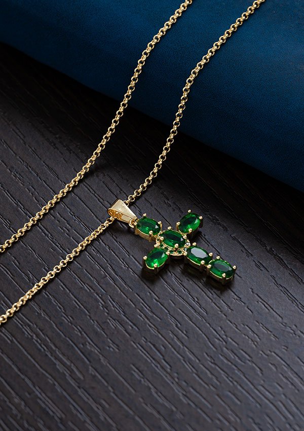 Un collar de oro con un colgante en forma de cruz adornado con piedras preciosas verdes se muestra elegantemente sobre una superficie oscura. Esta exquisita pieza es parte de nuestra colección Joyas en oro laminado 18k y se conoce como Cadena Aros 2 mm 65 cms.