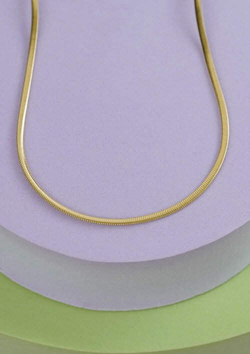 Vista de primer plano de un collar de oro, Cadena Serpiente 65 Cms elaborado con joyas en oro laminado 18k, colocadas sobre superficies superpuestas de colores pastel en tonos violeta claro y verde.