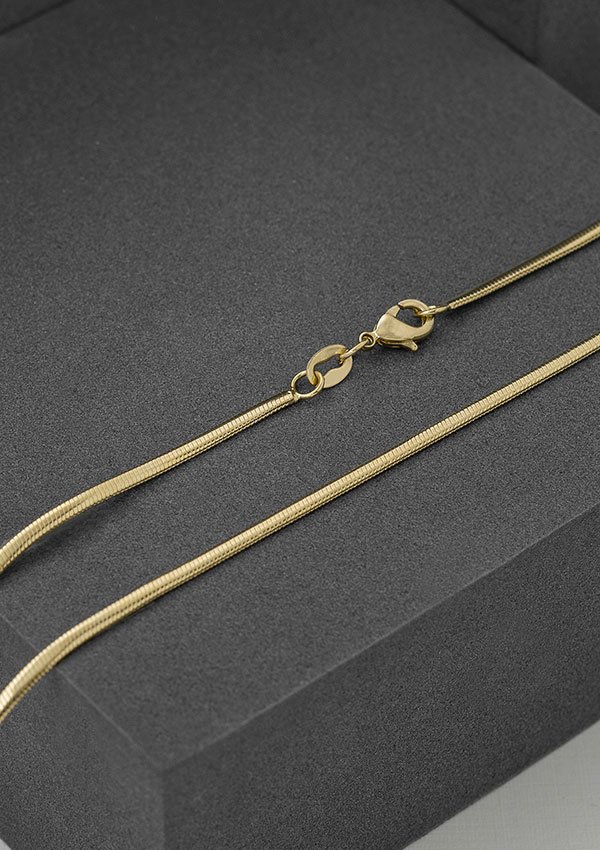 Cadena Serpiente 65 Cms, un collar de cadena de oro con cierre de langosta elaborado con joyas en oro laminado de 18k, está dispuesto sobre una superficie gris oscuro.