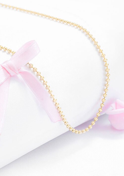 Un delicado collar de cuentas de oro, específicamente la Cadena Militar Mediana 65 Cms 2.5 Mm, reposa sobre una tela blanca con un lazo rosa y rosas, mostrando la belleza atemporal de las joyas en oro laminado de 18k.