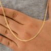 Una persona que lleva un collar de cadena de oro y una camisa gris coloca su mano sobre su pecho, mostrando su Cadena Cubana Plana Diamantada 65cm joyas en oro laminado 18k.