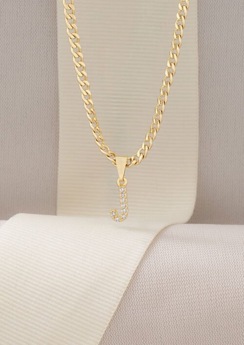 Un collar de cadena de oro llamado "Cadena Cubana 3 Mm 45cm" presenta un colgante con una letra "J" adornada con pequeños diamantes, sobre un lienzo de tela en tonos neutros, que encarna la elegancia de las joyas en oro laminado de 18k.