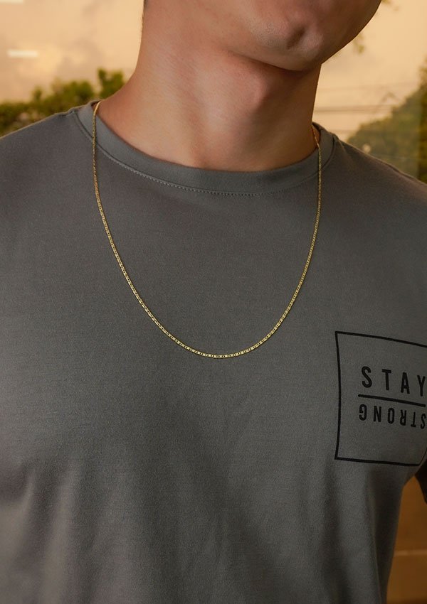 Primer plano de una persona que lleva una camiseta gris con el texto "STAY STRONG" en el bolsillo y un collar Cadena Escalera delg. Plana Lisa de 65 cm. No se ve el rostro de la persona.