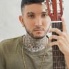 Una persona con tatuajes y pendientes sostiene una guitarra acústica, mirando directamente a la cámara, adornada con una Cadena Balin Discotequero e Italiano 65 Cms 4 mm.