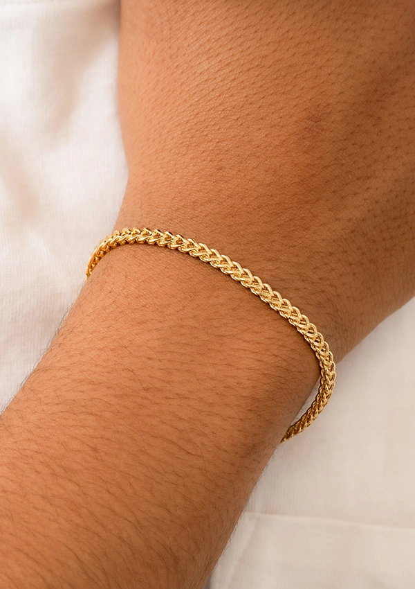 Una pulsera trenzada de oro, de la colección Joyas en Oro Laminado 18K, se usa alrededor de la muñeca de una persona, mostrada sobre un fondo blanco.