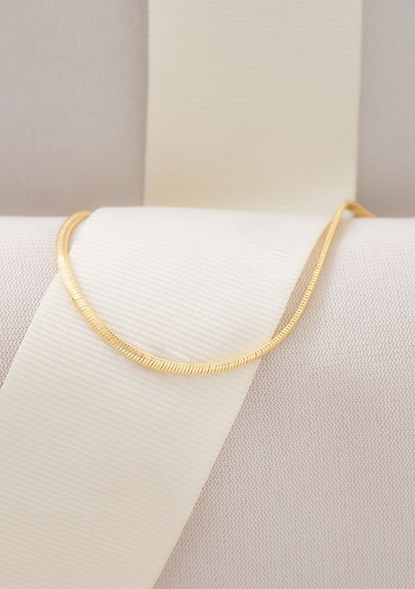 Una fina Tobillera Serpiente de oro de 25 cm expuesta sobre un fondo de tela blanca con una cinta vertical muestra la elegancia de las joyas en oro laminado de 18k.