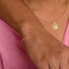 Una persona que lleva joyas de oro laminado de 18 quilates (una pulsera de oro, un collar de oro con un colgante de trébol y un anillo de oro) apoya su mano sobre el hombro de alguien. Está vestida con una blusa rosa.