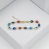 La Pulsera Ojo Turco Multicolor 16 Cms + Extensor en joyas en oro laminado 18k presenta dijes alternados en forma de ojos de color rojo, azul, celeste y negro que se muestran sobre un fondo geométrico blanco.