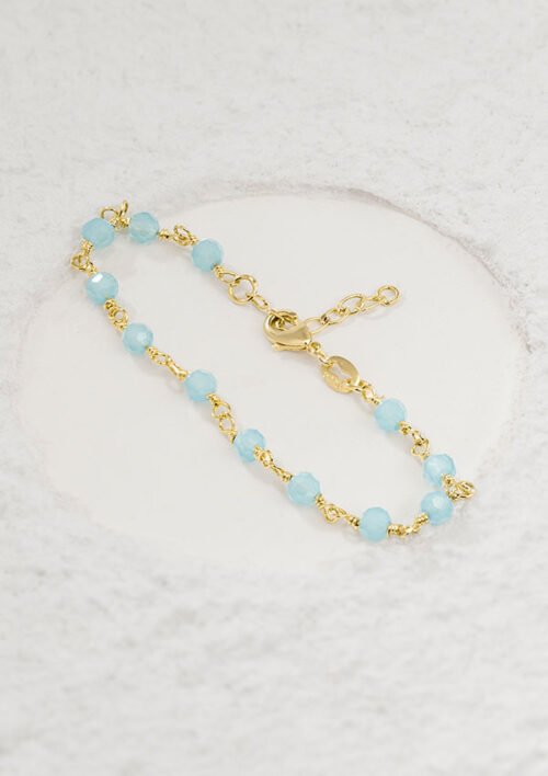 Una delicada pulsera de cadena dorada adornada con pequeñas cuentas de color azul claro colocadas sobre una superficie texturizada de color blanquecino, que muestra la elegancia de la Pulsera Blue Dream 16 Cms + Extensor.