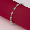 La Pulsera Blue Dream 16 Cms + Extensor, una pulsera de cadena de oro acentuada con cuentas de color azul claro y elaborada en joyas en oro laminado de 18k, se muestra elegantemente sobre una pieza enrollada de tela roja.