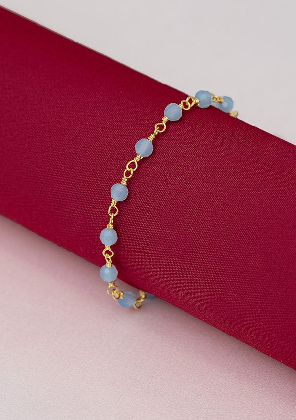 La Pulsera Blue Dream 16 Cms + Extensor, una pulsera de cadena de oro acentuada con cuentas de color azul claro y elaborada en joyas en oro laminado de 18k, se muestra elegantemente sobre una pieza enrollada de tela roja.