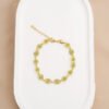 Una pulsera Sweet Lemon de 16 cm con piedras preciosas verdes dispuestas en un patrón alterno, completa con extensor, exhibida en una bandeja blanca ovalada: joyas en oro laminado de 18k que exudan elegancia atemporal.