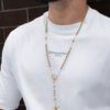 Primer plano de una persona que lleva una camisa blanca y un collar rosario de oro hecho con joyas en oro laminado de 18k contra una pared de ladrillos.