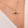 Primer plano de una persona luciendo una pieza de la colección Joyas en Oro Laminado 18K, mostrando un moderno collar de oro en forma de cruz con una pequeña piedra preciosa verde en el centro.