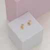 Dos pequeños aretes de Joyas en Oro Laminado 18K y perla se exhiben sobre una plataforma cúbica blanca con fondos cúbicos rosas y grises.