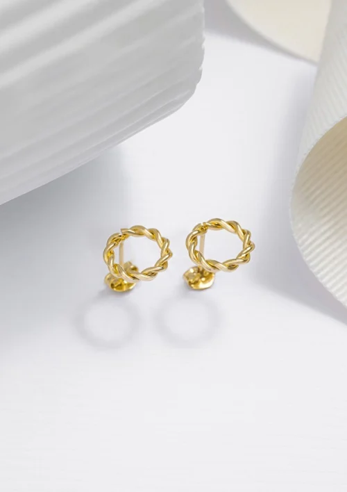 Un par de aretes de aro retorcidos bañados en oro de 18 quilates, etiquetados como Joyas en Oro Laminado 18K, colocados sobre una superficie blanca con cintas decorativas blancas parcialmente visibles en el fondo.