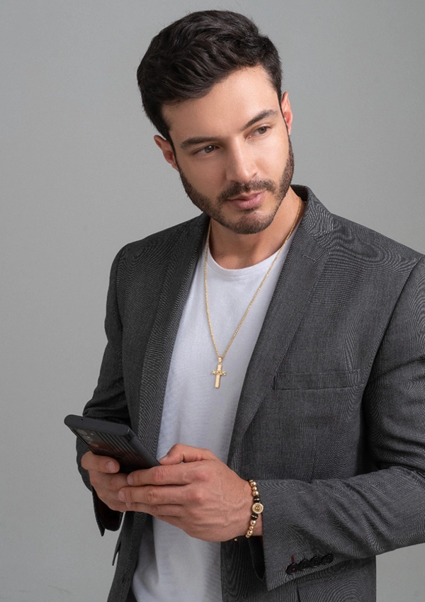 Un hombre de cabello corto y oscuro, vestido con un blazer gris, camisa blanca y un collar de cruz de oro de la colección Joyas en Oro Laminado 18K, mira hacia un lado mientras sostiene un teléfono inteligente en sus manos.