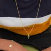 Se muestra a una persona que viste una camisa de rayas azul marino, blancas y amarillas de hombros hacia abajo, mostrando Joyas en Oro Laminado 18K: un collar de oro con una cruz verde y una pulsera de oro con un dije circular y una pequeña cruz.